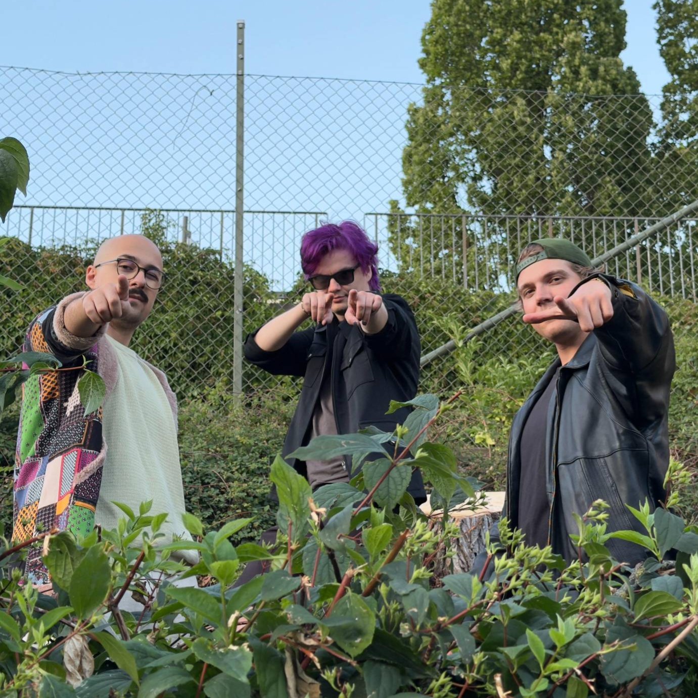 The three band members pointing at the camera through bushes